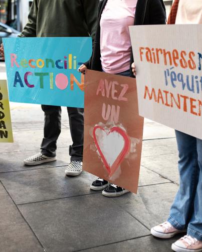 photo of children standing outside holding colourful hand painted signs reading, fairness now, l’équité maintenance, ayes un coeur, reconciliation and Jordan